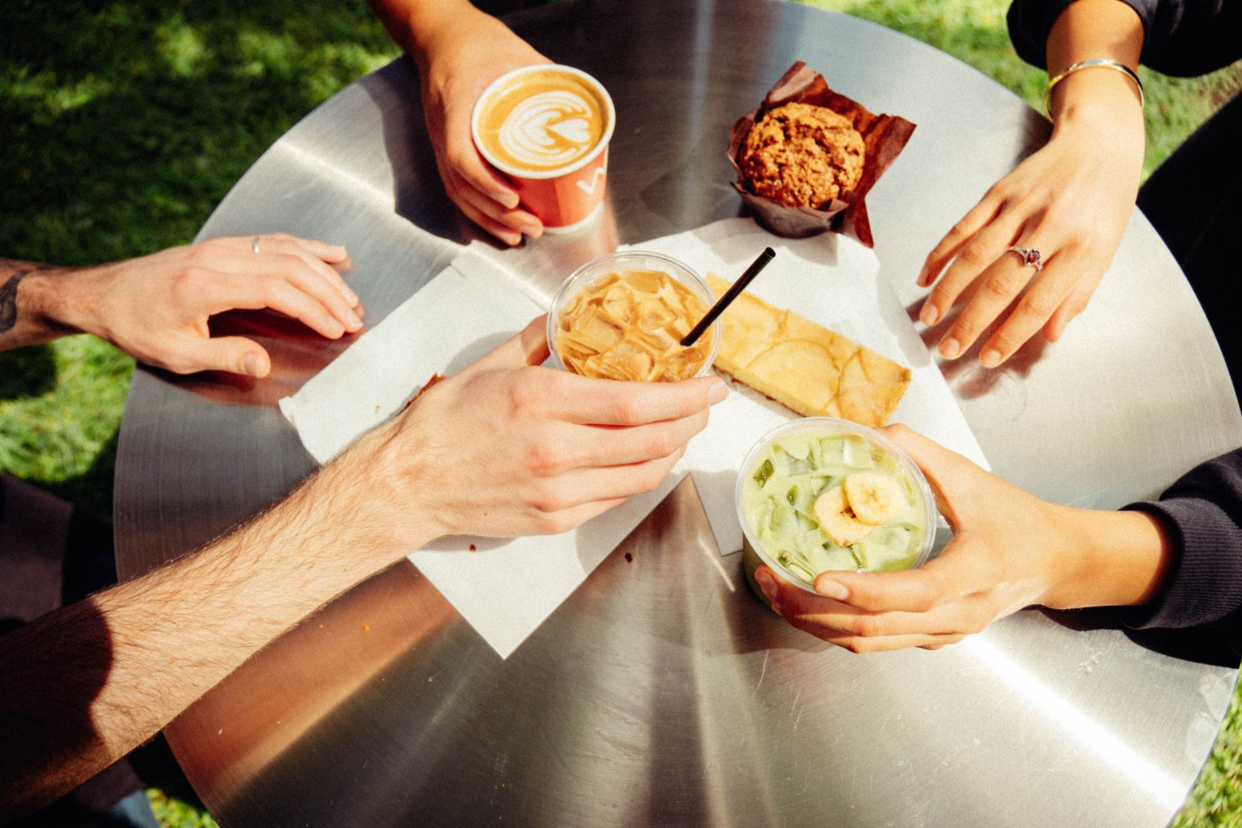 Friends sharing a hot latte, an iced coffee, an iced matcha with banana, and baked goods on a steel table outside